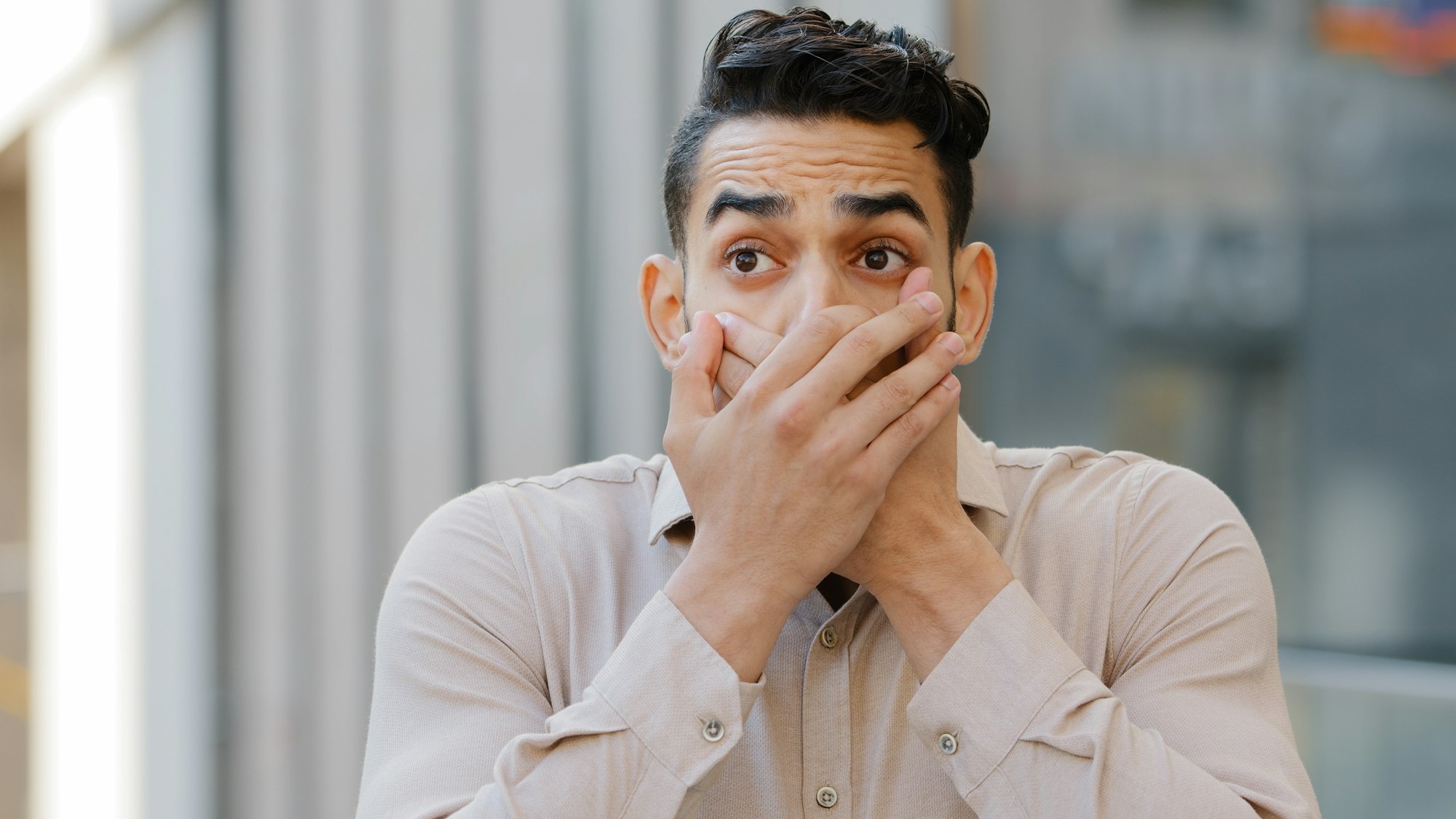 Hispanic man scared businessman boss stands in city covering mouth with hands from shock fear blab